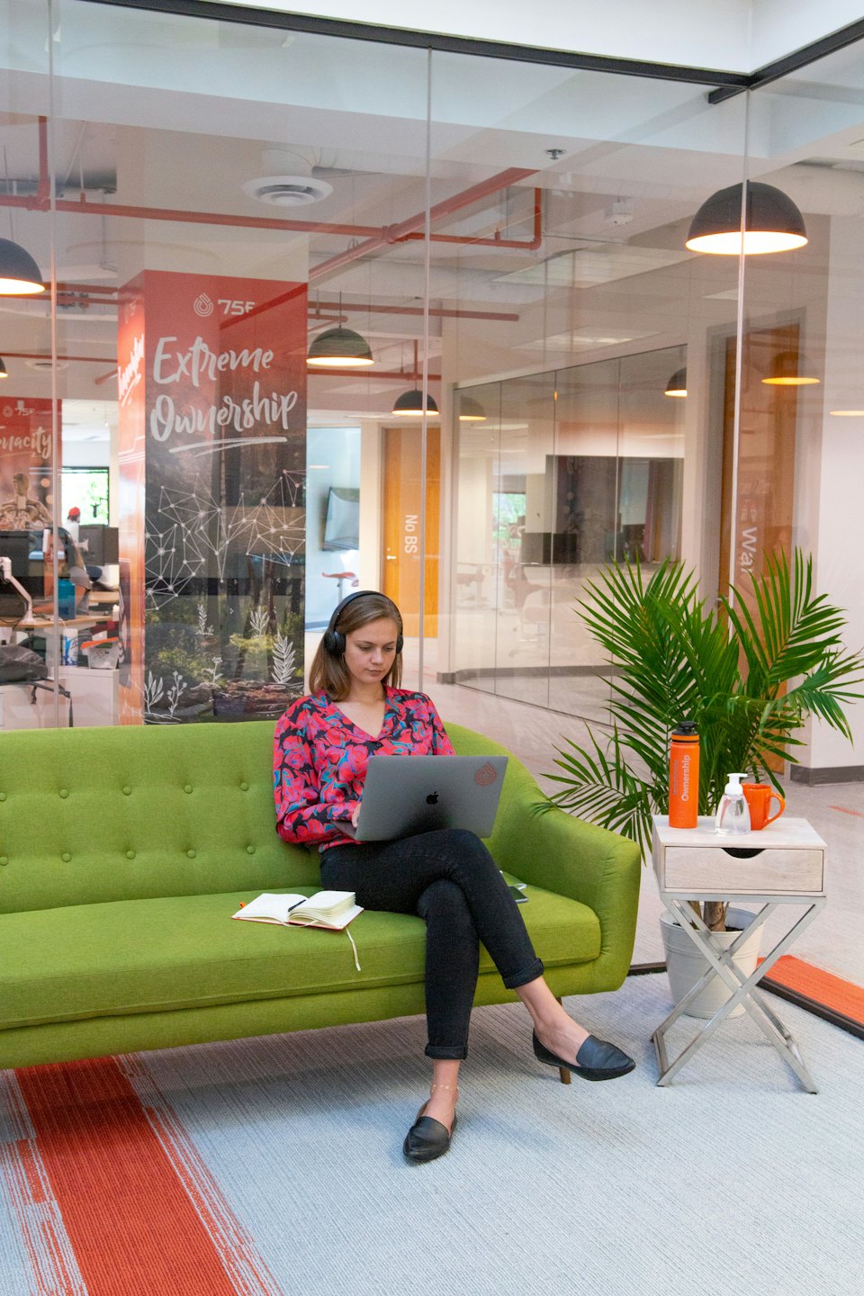 Woman working on laptop on a green sofa with headphones on.